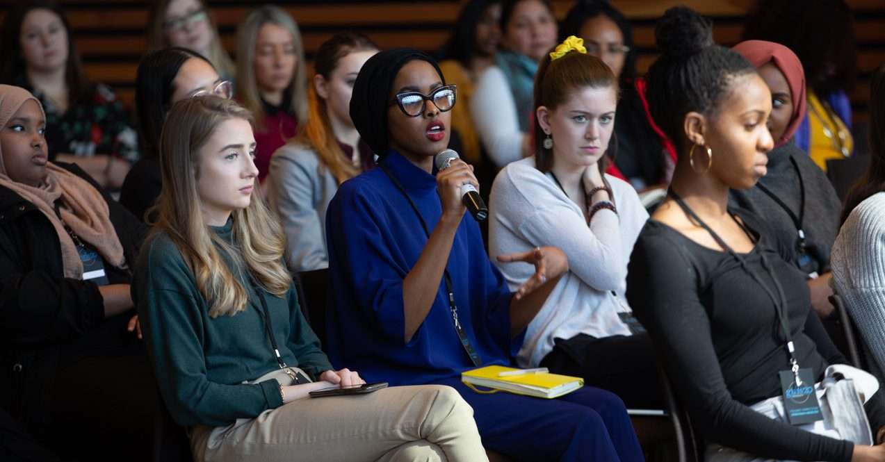 This is a photo of women sitting in an auditorium. The women in the middle is holding a microphone and speaking. She is wearing a royal blue outfit and big black glasses.