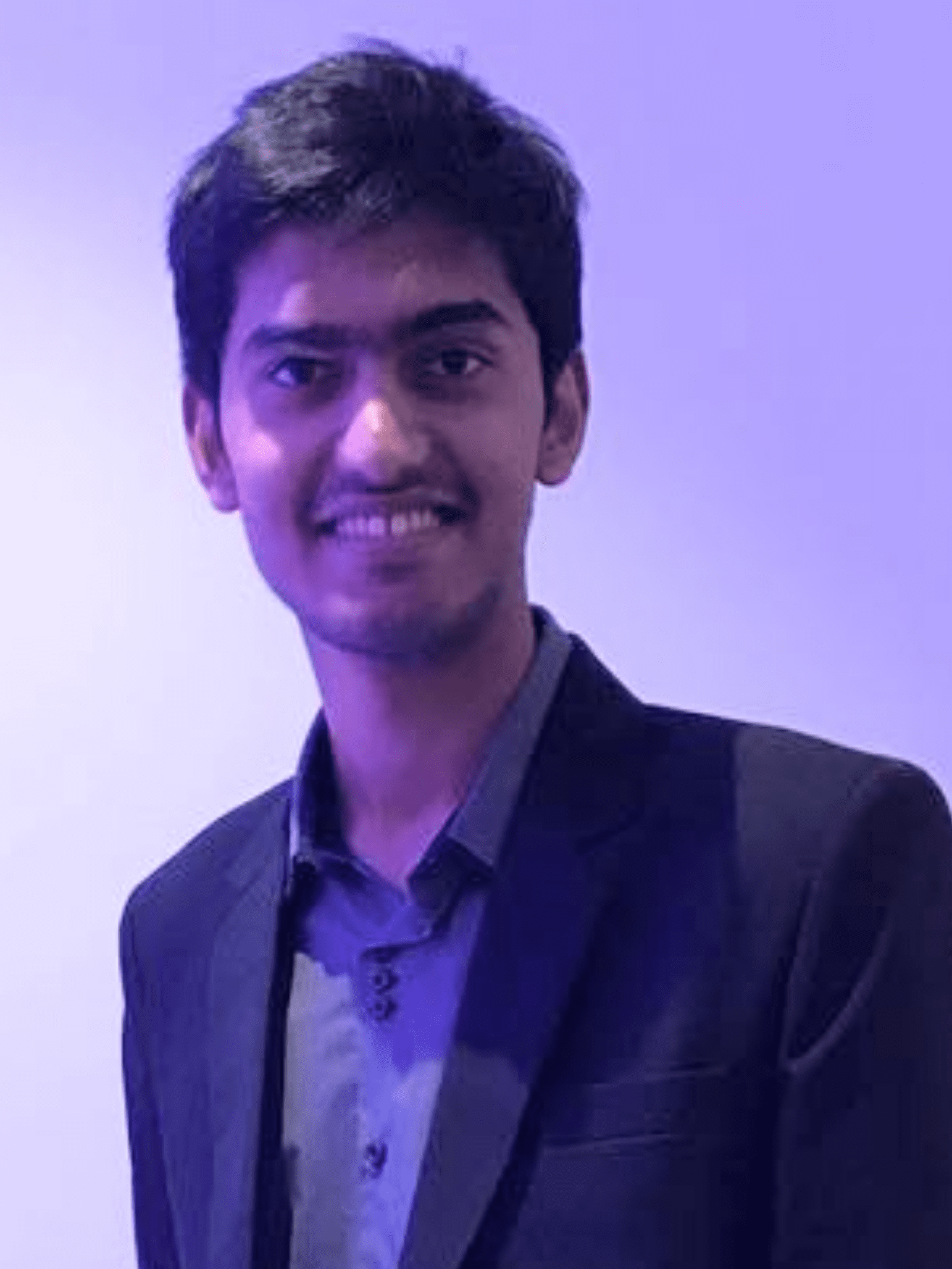 Rishit facing the camera with a smile wearing a black blazer and a grey shirt. Image has a plain background and a shade of purple because of the lights.