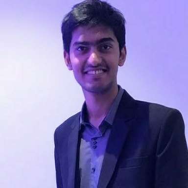 Rishit facing the camera with a smile wearing a black blazer and a grey shirt. Image has a plain background and a shade of purple because of the lights.