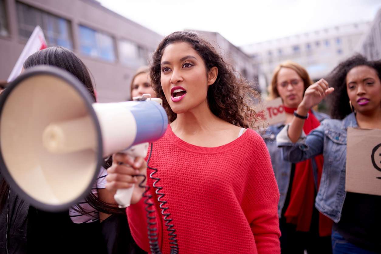 Woman in red shirt at a protest, holding a megaphone.