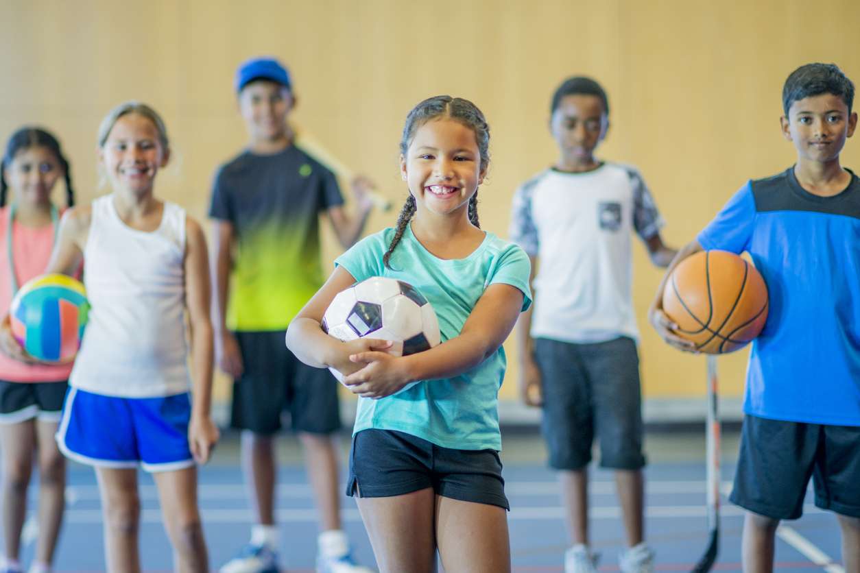Photograph of a group of smiling kids standing in a gymnasium. They are holding a basketball, volleyball, hockey stick, skipping rope, baseball bat, and soccer ball.